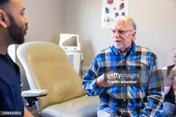 senior man speaking with doctor in the exam room with his wife in the background - biopsy stock pictures, royalty-free photos & images