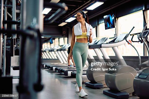 mujer en forma caminando en el gimnasio después de entrenar, sosteniendo una botella de agua y escuchando música - centro de bienestar fotografías e imágenes de stock
