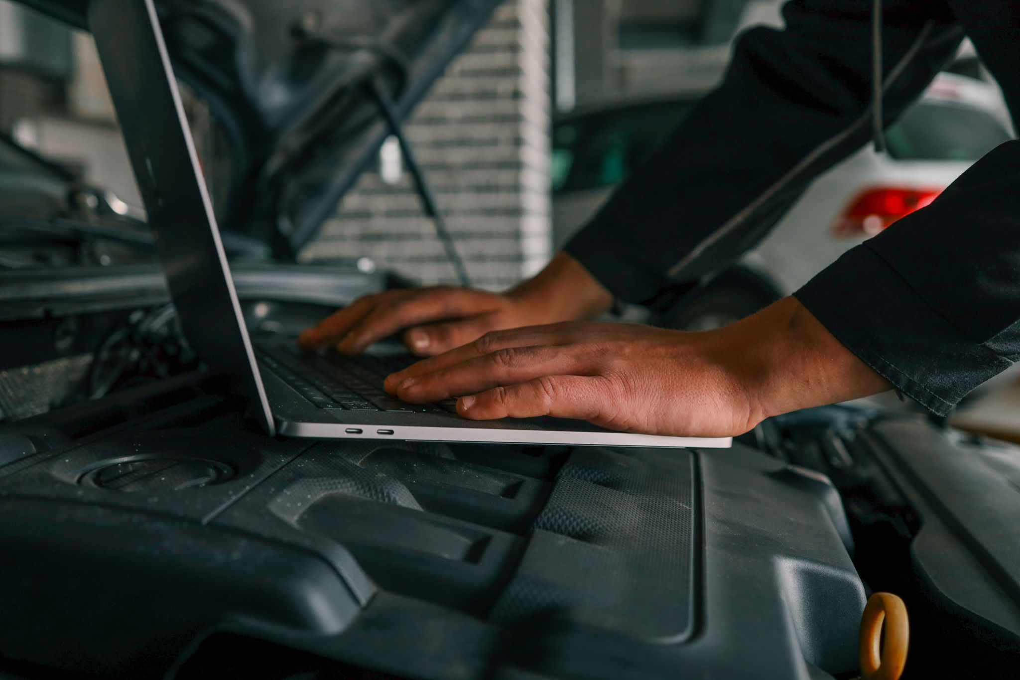 Close-up of a mechanic's hands typing on a laptop placed on a car engine in a workshop, showcasing the integration of technology in vehicle diagnostics and repairs Close-up of a mechanic's hands typing on a laptop placed on a car engine in a workshop, showcasing the integration of technology in vehicle diagnostics and repairs