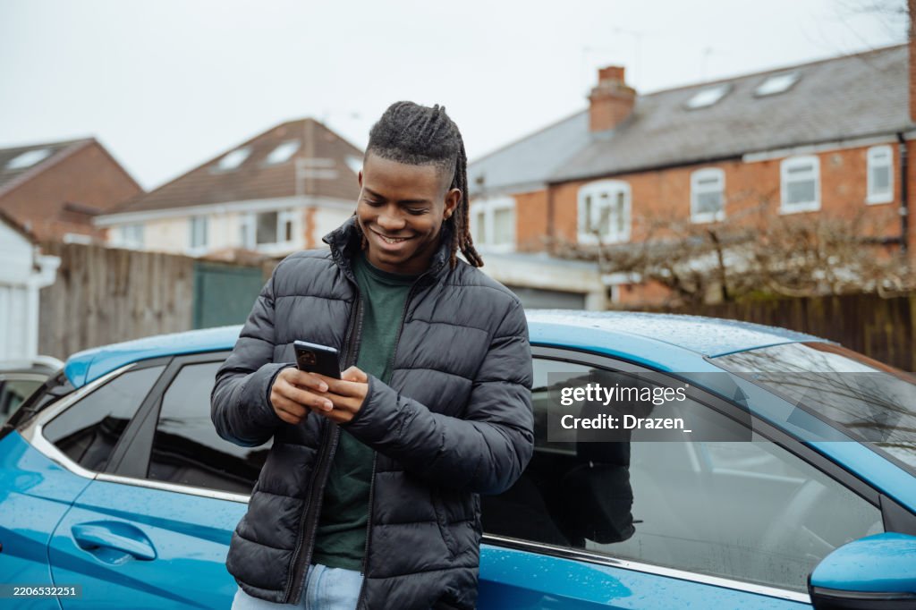 Teenage boy standing near the blue car and using smart phone for texting and voicemail