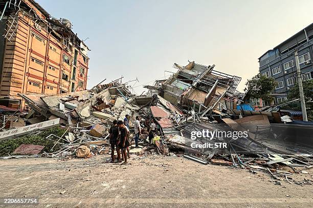 People stand past the debris of a collapsed building in Mandalay on March 28 after an earthquake. A powerful earthquake killed more than 20 people...