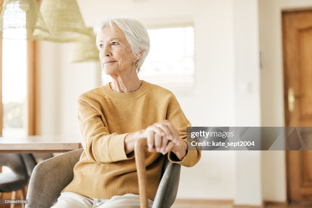 Ein Empowered-Aging-Konzept, das durch eine strahlende, weißhaarige Frau gezeigt wird, die in einer komfortablen häuslichen Umgebung lächelt.