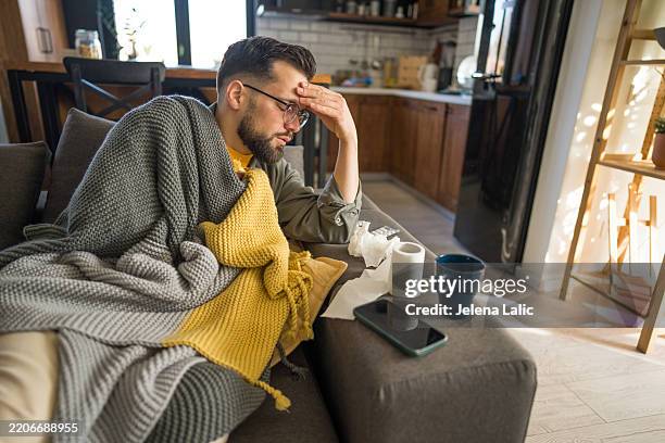 kranker junger mann, der an erkältung oder grippe leidet, sitzt auf einem sofa, das mit einer decke bedeckt ist - fieber stock-fotos und bilder