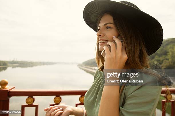 smiling young woman in an olive dress and a black wide brim hat talking on smartphone on a bridge - knob hill stock pictures, royalty-free photos & images