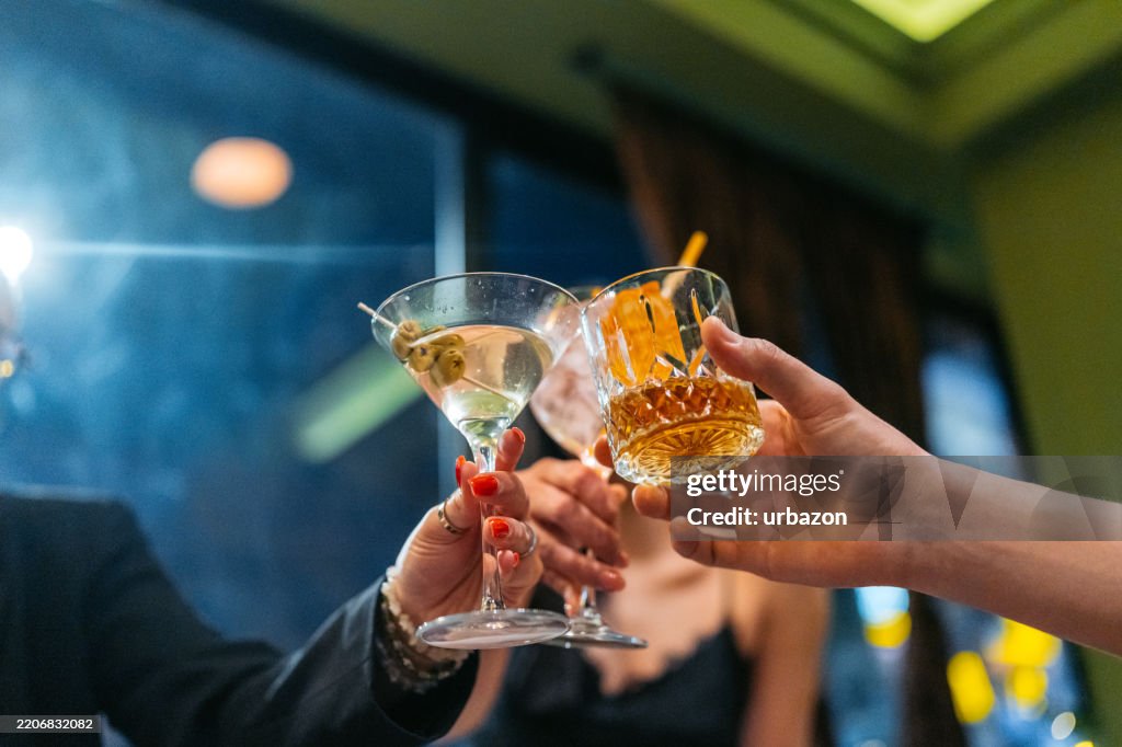 Three Young Friends Having A Celebratory Toast With Cocktails And Whiskey In A Bar At Night