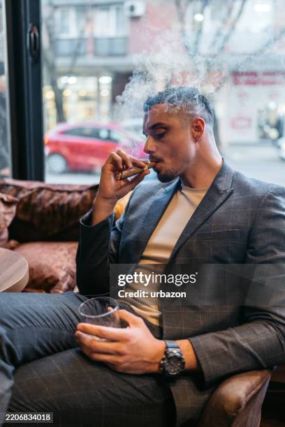 young man smoking a cigar while drinking whiskey in a bar - whiskey tasting stock pictures, royalty-free photos & images