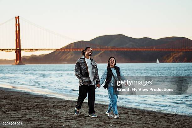couple heureux marchant sur la plage de san francisco au coucher du soleil - région de la baie de san francisco photos et images de collection