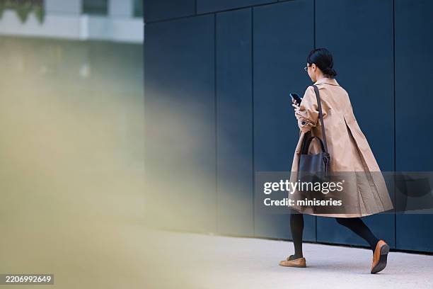 businesswoman walking in the city while using her smartphone - long coat stock pictures, royalty-free photos & images