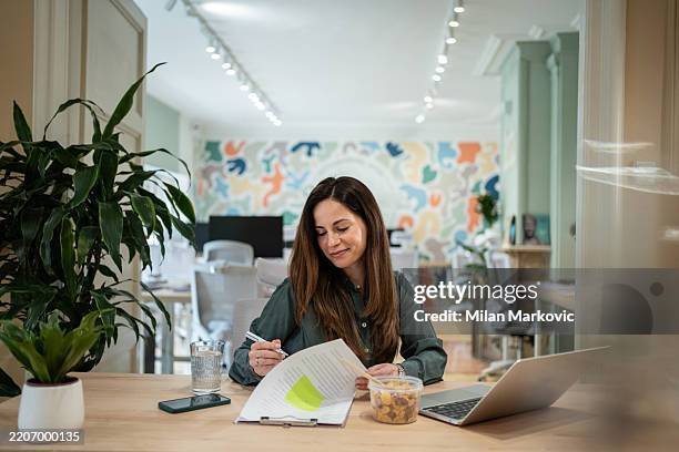 businesswoman reviewing documents and having a snack at her desk in a modern office - cfo stock pictures, royalty-free photos & images