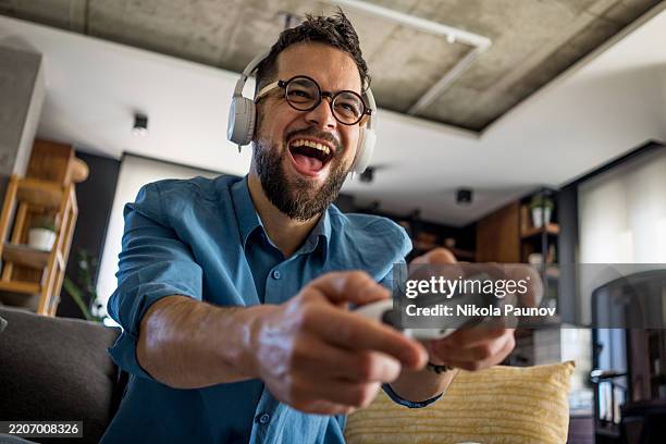 a man in a relaxing living room is focused on playing a video game with a joystick and headphones on his head. his face reflects joy and concentration. the comfortable sofa and modern interior create the perfect atmosphere for relaxation and fun at home - videospel stockfoto's en -beelden