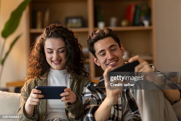 couple heureux jouant à des jeux mobiles sur des smartphones tout en étant assis sur le canapé à la maison - jeu mobile photos et images de collection
