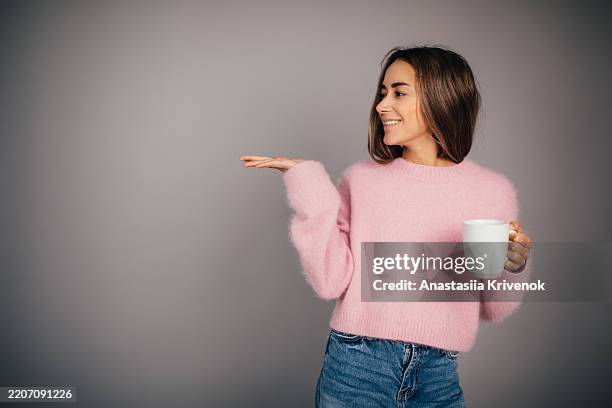 smiling woman holding a mug and gesturing against a studio background - positioning stock pictures, royalty-free photos & images