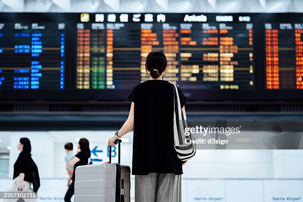 rear view of young asian woman carrying suitcase, looking up and checking flight schedule from the arrival departure board at airport terminal. female traveller ready for a trip. businesswoman on business travel. travel and vacation concept - train billboard stock pictures, royalty-free photos & images