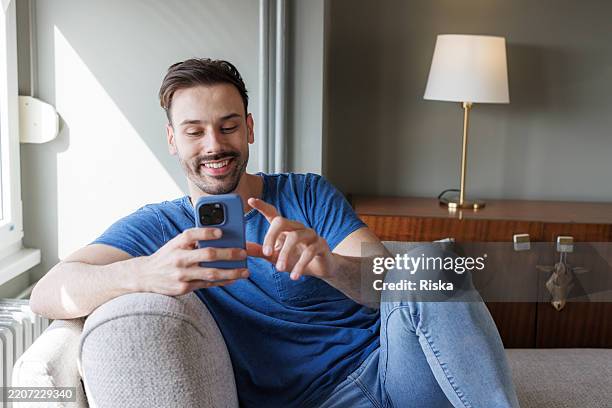 smiling man relaxing on sofa while using smart phone at home - blue couch stock pictures, royalty-free photos & images