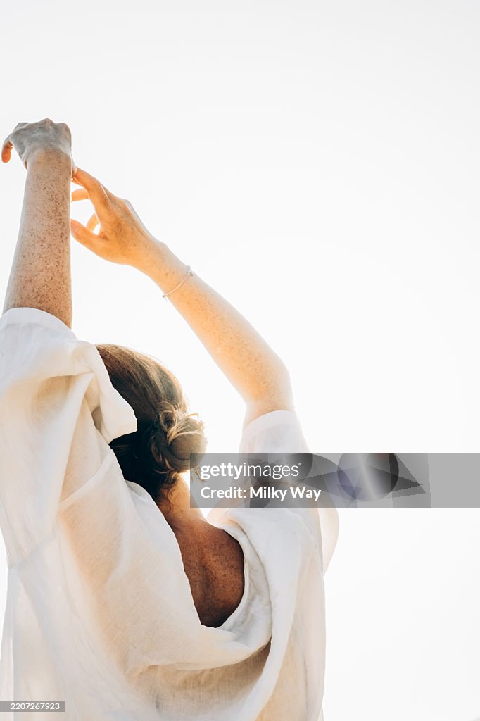Serene morning stretch: tranquil woman in sunlit atmosphere. Red-haired woman in white loose clothing stretches her arms upward. Bright backlight creates minimal, high-key style.