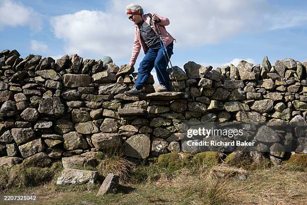 Woman walker climbs over a dry stone wall stile in the Lake District National Park, on 24th March 2025, in Keswick, Cumbria, England.