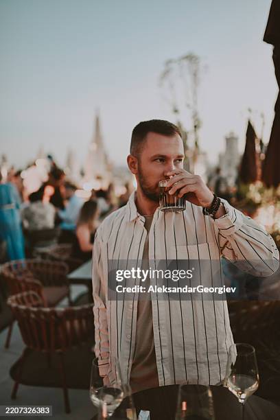 glücklicher junger mann, der sich in seiner rooftop-bar einen whisky gönnt und den sonnenuntergang genießt - whisky stock-fotos und bilder