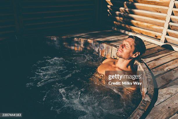 mature man with short relaxes in a bubbling wooden hot tub. eyes closed, face in sunlight. warm tones and shadows. hydrotherapy, wellness and retreat lifestyle concept. - kuuroord stockfoto's en -beelden