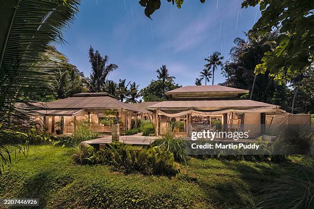 tented villa at eco friendly luxury glamping resort with open side panels showing open plan interior, situated amongst lush tropical foliage outdoors, shot during full moon, long exposure with star trails. - safari lodge stock pictures, royalty-free photos & images