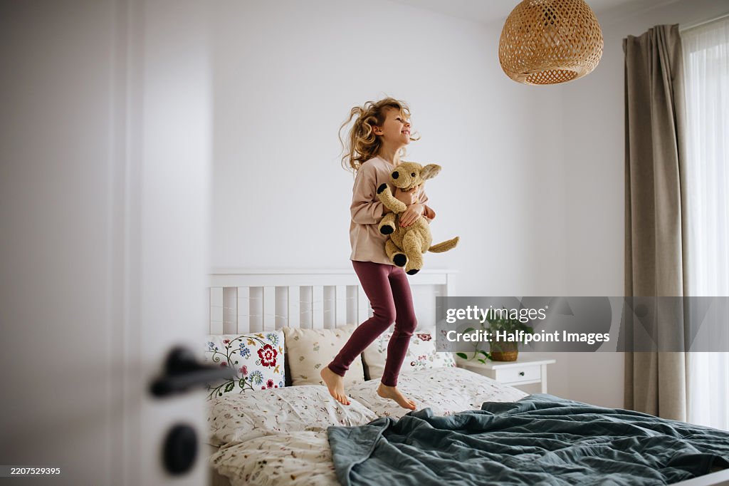 Cute young girl jumping on bed with plush toy in arms.