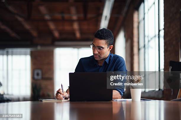 focused professional writing notes while working on laptop in stylish office space - electronic organiser stock pictures, royalty-free photos & images