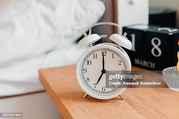 classic white alarm clock on wooden bedside table with morning light - bedside table stock pictures, royalty-free photos & images