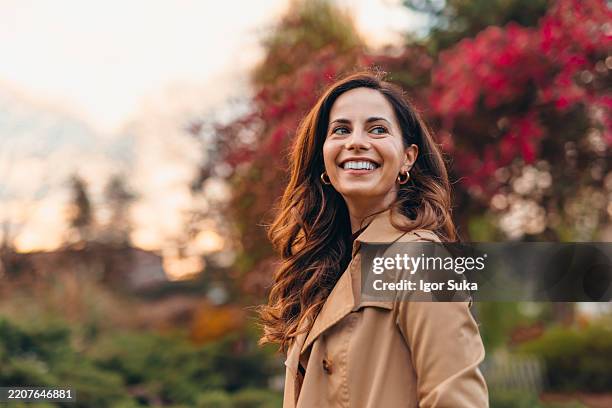 happy woman smiling and looking away in autumn park during sunset - trenchcoat stockfoto's en -beelden