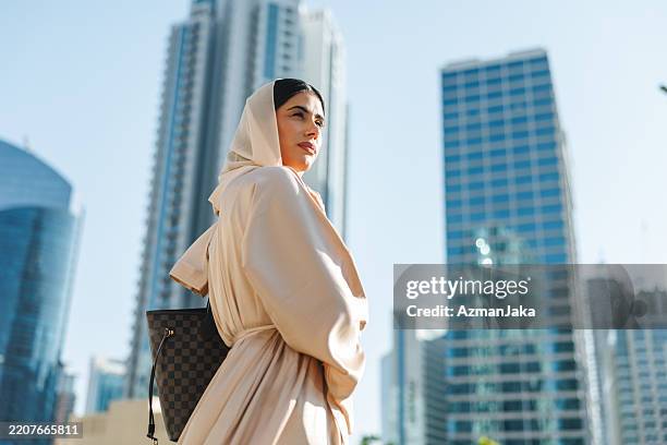 junge frau in cremefarbener abaya steht vor modernen wolkenkratzern in dubai - vereinigte arabische emirate stock-fotos und bilder