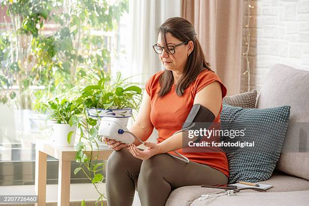 porträt einer frau, die mit einem blutdruckmessgerät auf einem sofa sitzt - bluthochdruck stock-fotos und bilder