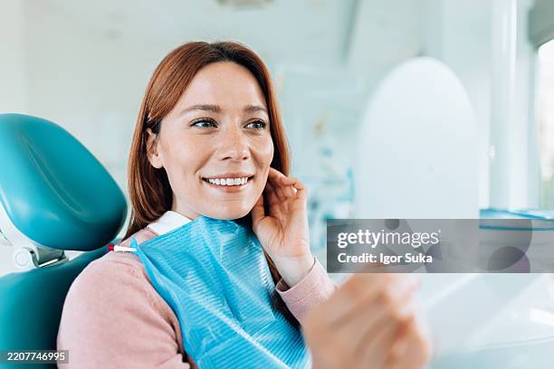 young woman smiling after dental checkup, admiring her teeth in the mirror - white teeth stock pictures, royalty-free photos & images