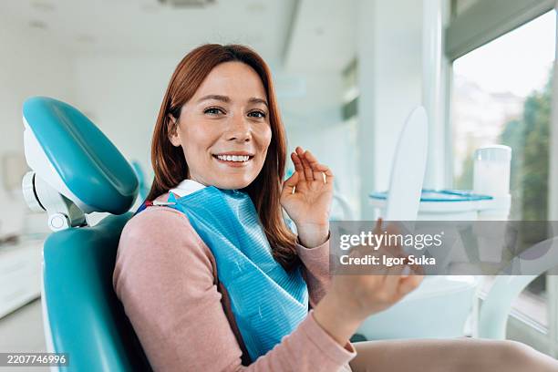 junge frau, die ihr lächeln nach der zahnärztlichen untersuchung in der zahnarztpraxis bewundert - dentalimplantat stock-fotos und bilder