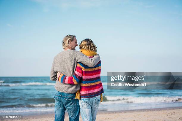 ture couple standing on the beach with arms around, looking at the sea - beschützer stock-fotos und bilder