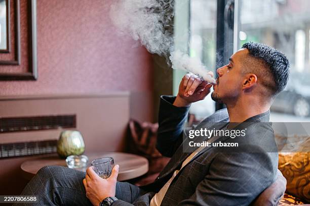 young man smoking a cigar while drinking whiskey in a bar - smokey bar stock pictures, royalty-free photos & images