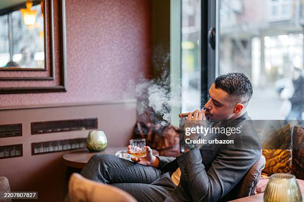 young man smoking a cigar while drinking whiskey in a bar - cigar bar stock pictures, royalty-free photos & images