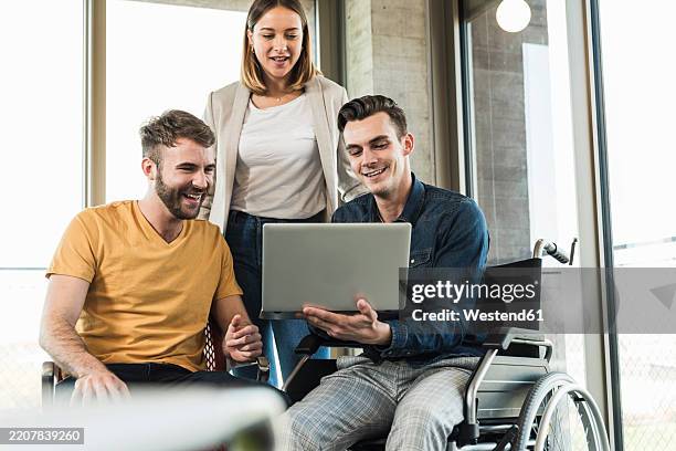 young businessman in wheelchair showing laptop to colleagues in office - chancengleichheit stock-fotos und bilder