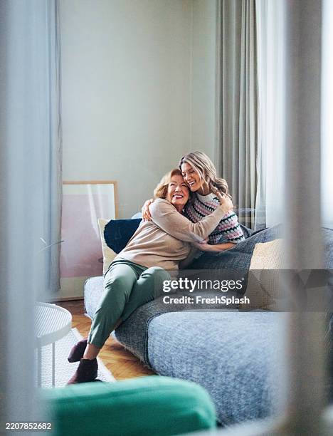 two women hugging and smiling happily on a comfortable living room sofa - tears of joy stock pictures, royalty-free photos & images