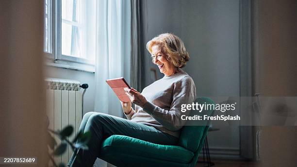 smiling elderly woman reading on a tablet by natural light indoors - brightly lit stock pictures, royalty-free photos & images