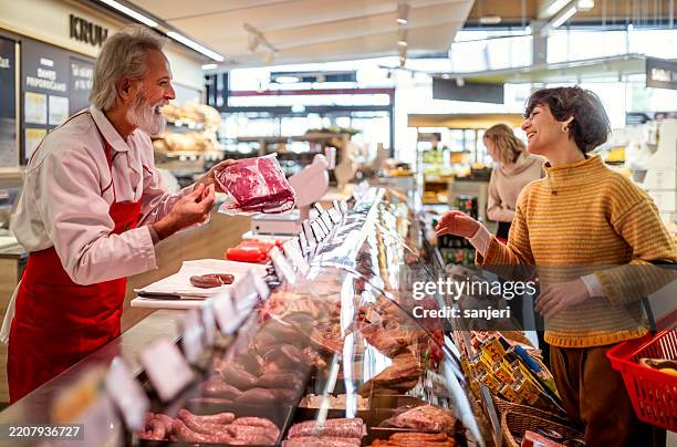 mature male shop assistant and smiling female customer in well-lit grocery store - butcher stock pictures, royalty-free photos & images