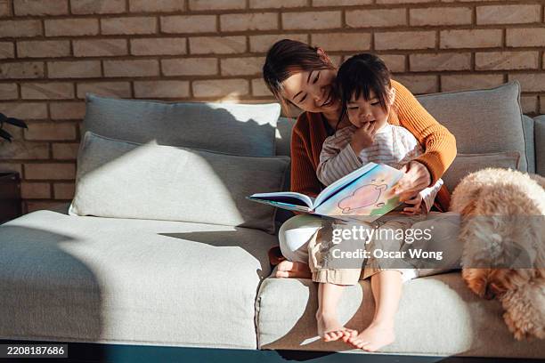 young asian mother reading picture book for cute daughter while sitting with their family dog on sofa at home - historia bildbanksfoton och bilder
