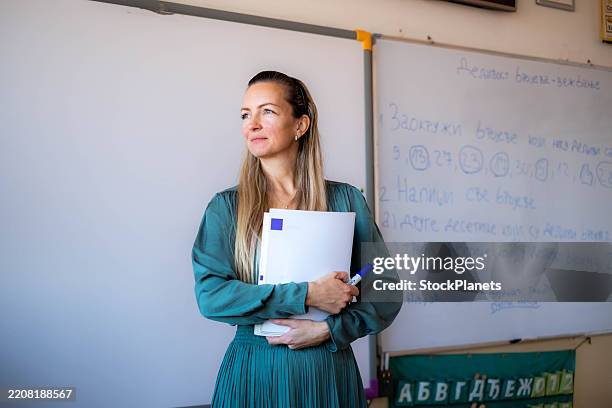 happy female teacher, teacher at school standing in front of the blackboard and holding books in her hands - instructor stock pictures, royalty-free photos & images