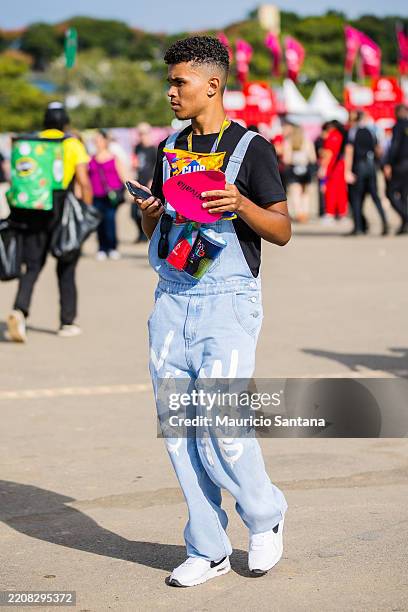 Festival-goer wears black crew neck t-shirt, light blue denim overalls with white graphic print, white athletic sneakers with black details, festival...