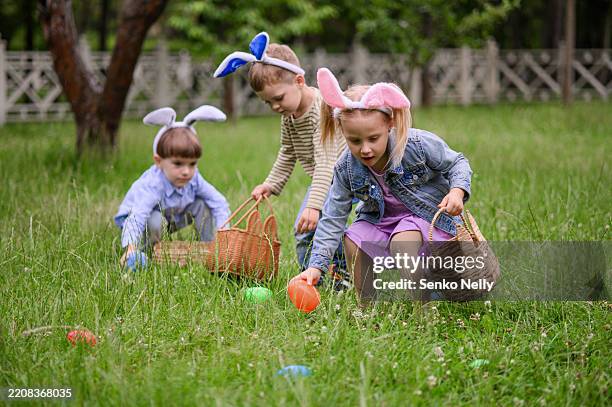 children wearing bunny ears collecting easter eggs outdoors in spring - easter egg hunt stock pictures, royalty-free photos & images