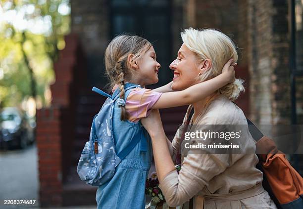 mother embracing daughter ready for school outdoors on a sunny day - primary age child stock pictures, royalty-free photos & images