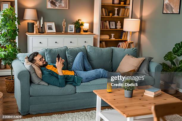 happy young man relaxing on sofa and using smart phone at home. - ligga ner bildbanksfoton och bilder