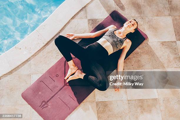 relaxed woman meditating outdoors by poolside yoga mat in sunlight. young woman lies in reclined butterfly pose on yoga mat next to swimming pool. her eyes are closed and face relaxed. peaceful atmosphere with sunlit tiles and soft reflections in water. - colchoneta de ejercicio fotografías e imágenes de stock