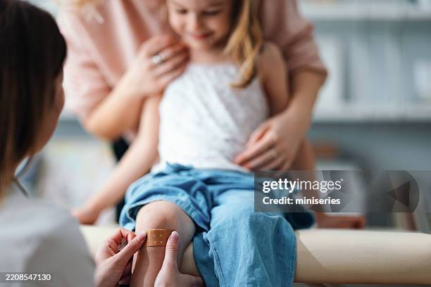 mother comforts daughter with doctor applying bandage to injured knee - bandage stock pictures, royalty-free photos & images