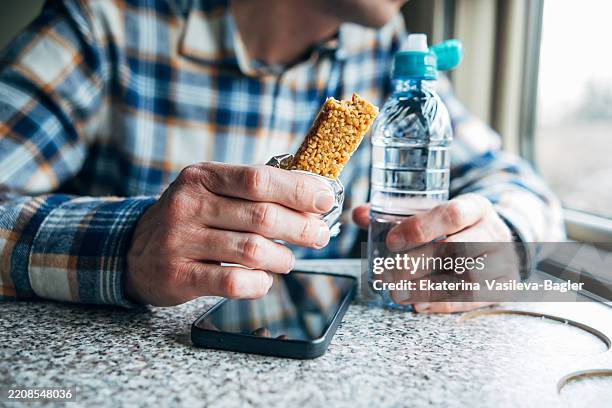 healthy snack on the go, man's hand holding cereal bar and bottle of water on train during trip - muesli bar stock pictures, royalty-free photos & images