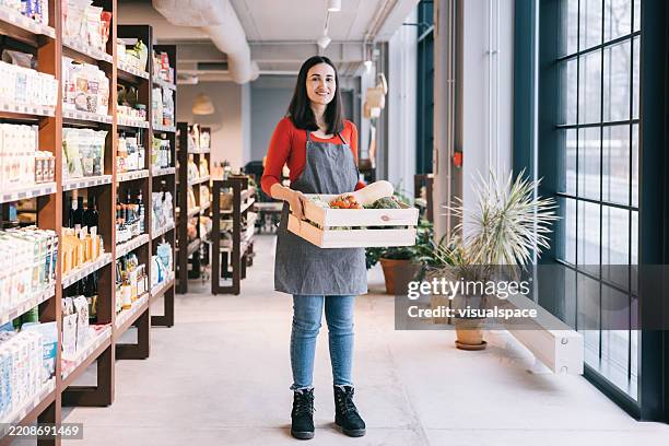 a smiling female latin shopkeeper stands inside the store, holding a box filled with fresh fruits and vegetables - duurzaam consumeren stockfoto's en -beelden