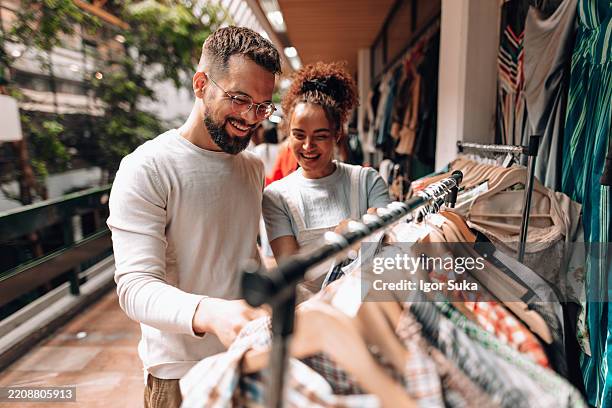 young couple searching clothes in a vintage second hand shop - mid adult couple stock pictures, royalty-free photos & images