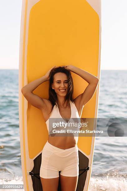 side view portrait of a mixed woman standing in the surf with a surfboard under her arm. the mid adult woman in a swimsuit spends her weekends on the beach on vacation at sea sup boarding on the water - beauty in nature stock pictures, royalty-free photos & images